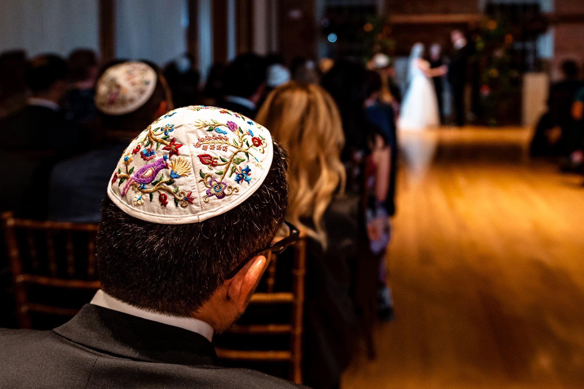 photo shows the back of a man's head as he watches a Jewish wedding ceremony. he is wearing an embroidered yarmulke | photo by Kivus & Camera