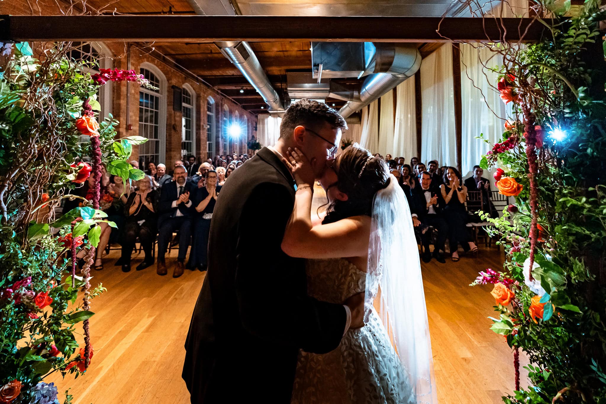 couple shares their first kiss under a floral adorned chuppah at the Cotton Room in Durham, NC | Photo by Kivus & Camera